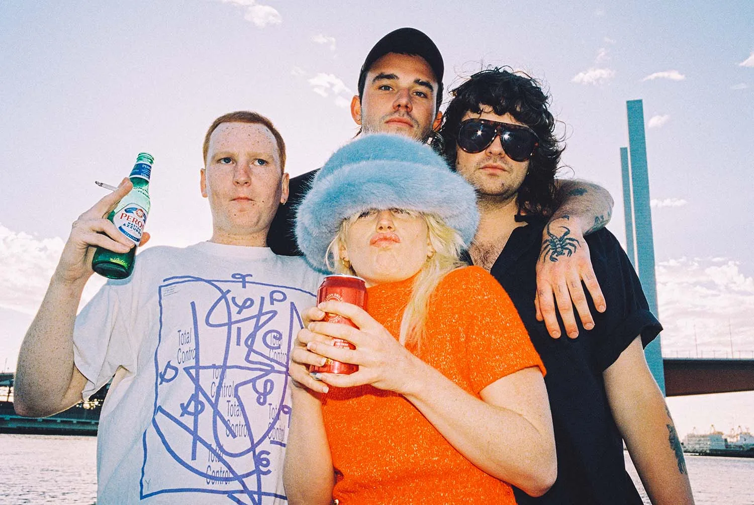 Amyl and the Sniffers pose together outdoors in a bright press portrait, with Amy Taylor centered in a blue hat.
