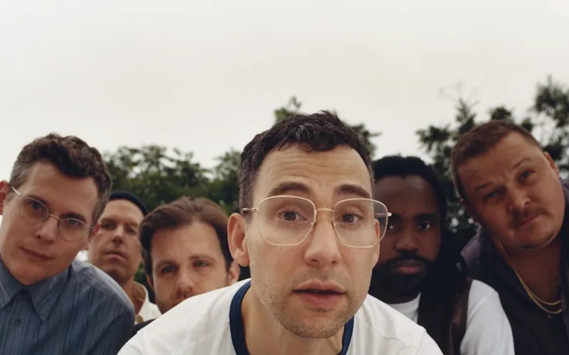 Jack Antonoff leans toward the camera with members of Bleachers gathered around him outdoors.