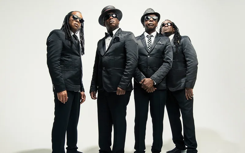 The four members of Living Colour pose together in black suits and sunglasses against a light studio backdrop.
