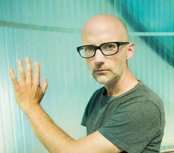 Moby poses beside a frosted glass wall wearing black glasses and a gray T-shirt.