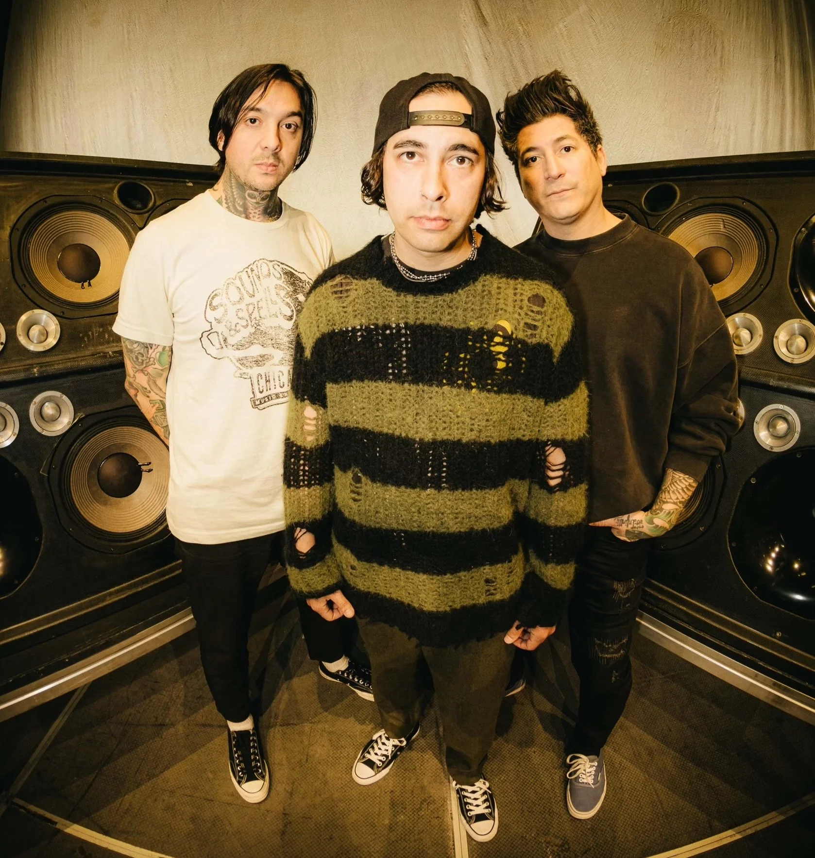 Pierce the Veil standing together in front of a wall of studio speakers in a promotional band photograph.