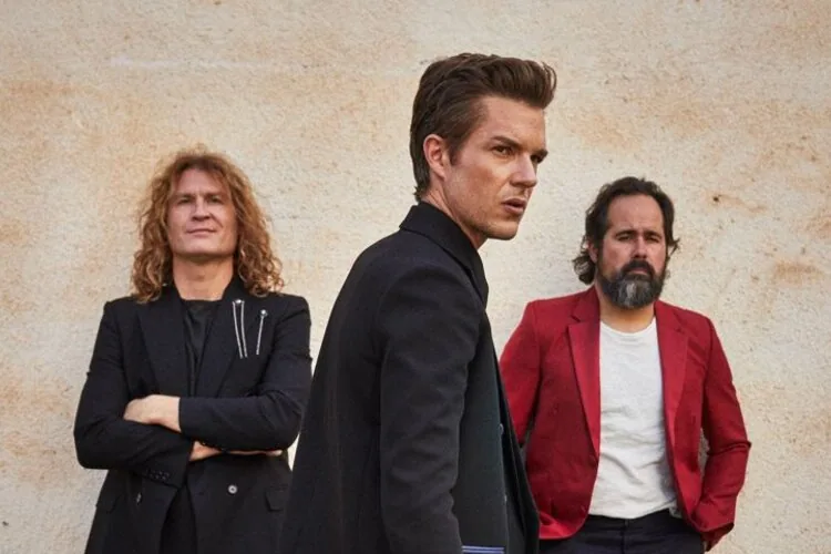 Members of The Killers pose in front of a beige wall, with Brandon Flowers centered in a dark suit.