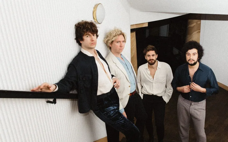 The Kooks pose together indoors in a modern curved hallway, wearing relaxed suits and open-collar shirts.